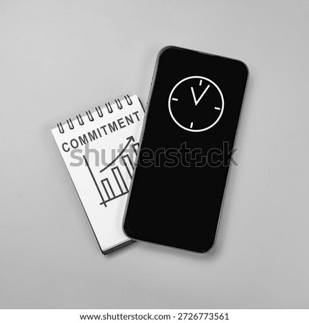 Top view flat lay of a notebook with 'COMMITMENT' and a growth graph under a smartphone displaying a clock icon, representing time management and business success