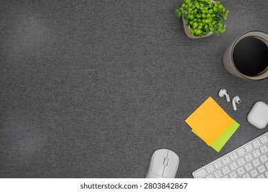 Top view flat lay of office workspace essentials including a clipboard, spiral notebook, pencil, sticky notes, coffee cup, and green plant on a grey fabric background with copy space