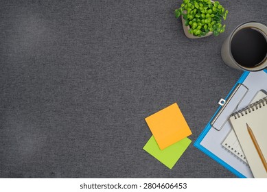 Top view flat lay of office workspace essentials including a clipboard, spiral notebook, pencil, sticky notes, coffee cup, and green plant on a grey fabric background with copy space