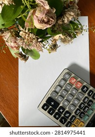 Top view flat lay of an office desk with blank white paper, calculator, and a bouquet of dry withered flowers on a wooden table