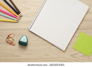Top view flat lay of open blank spiral notebook with grid graph paper pages, sharpened colorful wooden pencils, pencil sharpener with shavings, and green sticky note placed on light wooden background, creative education layout with copy space for back to school concept.