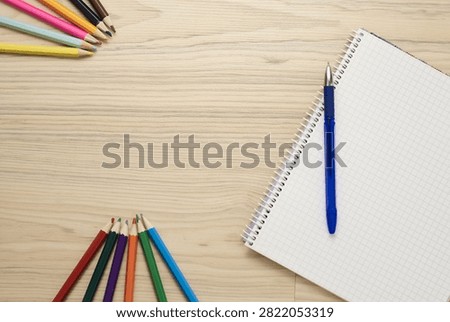 Top view flat lay of open blank spiral notebook with white grid graph paper pages and blue ballpoint pen on top, arranged with colorful wooden pencils over light natural wooden desk surface, minimal educational layout with copy space for back to school concept.