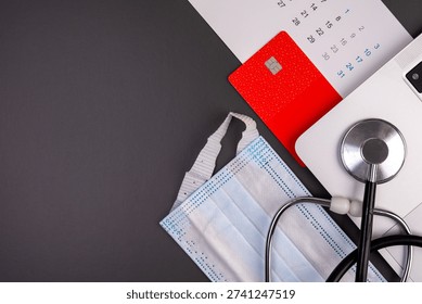 Top view flat lay of a red credit card on a calendar with a laptop, stethoscope, and surgical mask on a dark background. Concept of booking private medical appointments online, health insurance premium deadlines, and healthcare costs. Copy space.
