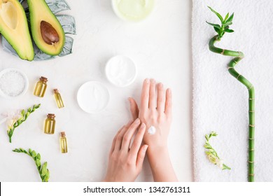 Top view and flat lay of woman puts cream on hands over white table with cosmetic products - avocado oil, cream and bamboo. Bod ycare concept