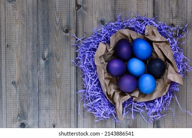 Top view flat lay of a wooden old rustic style table on which is placed a homemade nest made of paper shavings and craft paper, in which are painted blue and very peri festive Easter eggs, copy space. - Powered by Shutterstock