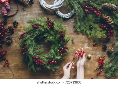 top view of florist hands making Christmas wreath with fir branches and decorative berries on wooden tabletop - Shutterstock ID 725913838