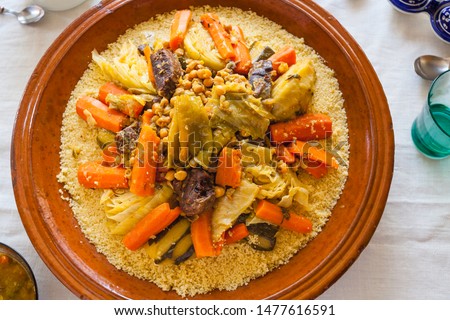 Top view food shot of a typical moroccan couscous served on a family size ceramics tagine plate surrounded by glasses, sauce, spoons and spices on a fine linens background. Blessed friday concept.