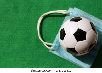 Top View Of Football Or Soccer Ball With Blurred Medical Face Mask On Green Background. The Concept Of New Normal For Sporting Event And Football Competition During Covid-19 Or Coronavirus. 