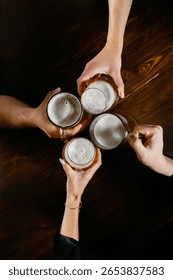 Top view of four hands clinking beer mugs over a dark wooden table. Foamy light beer in clear glass mugs. Two men and two women participating in a celebratory toast.