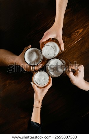 Top view of four hands clinking beer mugs over a dark wooden table. Foamy light beer in clear glass mugs. Two men and two women participating in a celebratory toast.