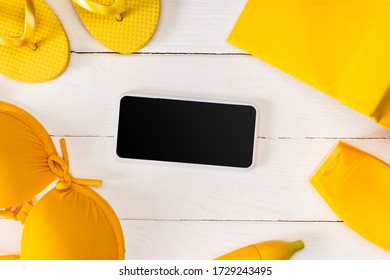 Top View Of Frame From Yellow Flip Flops With Sunscreen, Swimsuit And Book Near Smartphone On White Wooden Surface