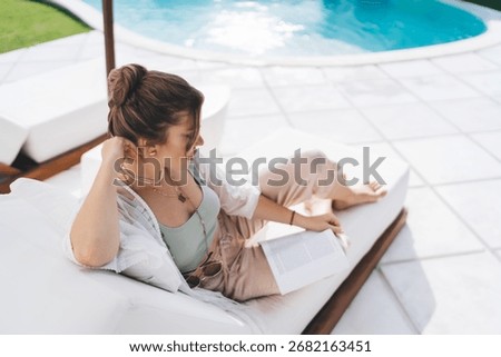 Top view of freelancer reading from notebook, relaxing on daybed near pool. Minimalism, independence, and deep focus define her creative break in tropical paradise.