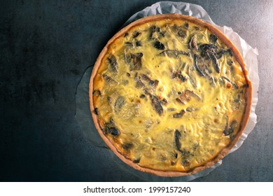 Top View Of A Freshly Baked Yellow Quiche With Mushrooms Still On A Baking Paper, Sitting On A Dark Kitchen Table Surface.