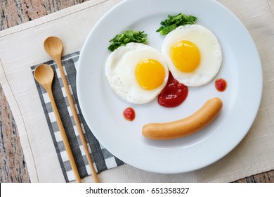 Top View Of Fried Egg Smile Face On White Plate With Spoon Knife And Fork On Purple Fabric Table, Breakfast Serve For Kid