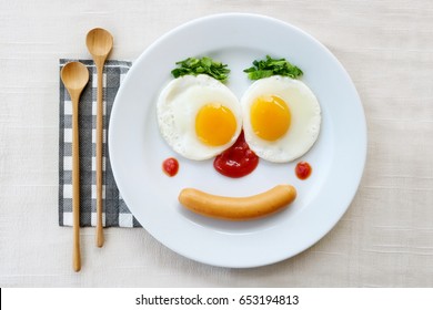 Top View Of Fried Egg Smile Face On White Plate With Wooden Spoon On White Fabric Table, Breakfast Serve For Kid