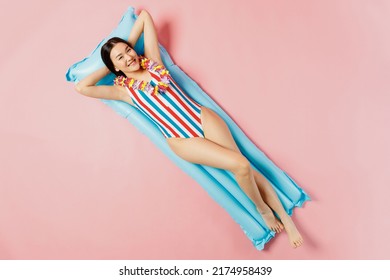 Top view full body fun woman of Asian ethnicity in striped swimsuit hawaii lei lies on inflatable mattress hotel pool isolated on plain pastel pink background. Summer vacation sea rest sun tan concept