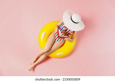 Top view full body young woman in striped swimsuit lies on inflatable rubber ring pool read book cover face with hat isolated on plain pastel pink background. Summer vacation sea rest sun tan concept