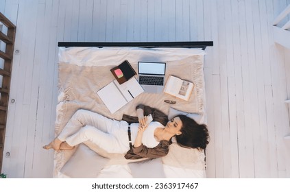 Top view full length of happy young woman in white top and trousers lying on bed and texting message via cellphone during break from remote studies on laptop - Powered by Shutterstock