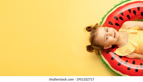 Top view of funny little child girl dressed in a swimsuit lying on an inflatable color red ring on yellow studio background. Summer travel concept. Banner, mock up, copy space