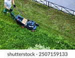 Top view of gardener in uniform using lawnmower mowing lawn in garden. Worker male working gardening machine tools, process of cutting grass. Gardening and landscaping concept. Copy ad text space