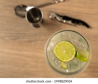 Top View Of Gin And Tonic Cocktail With Lemon Slice On Wooden Surface With Mixing Spoon, Jigger And Bottle Opener Out Of Focus.