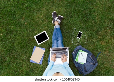 Top view of girl sitting in park on the green grass with laptop, tablet, smartphone with earphones and backpack. Copy space on screens. Flat lay