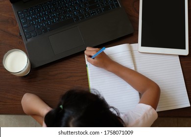 Top View Of A Girl Writing The Book With Tablet, Notebook, A Glass Of Milk On The Table. Children And Technology, Healthy Food For Kid Concept.