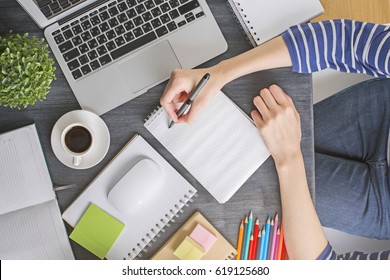 Top View Of Girl's Hand Writing In Empty Notepad Placed On Office Desktop With Laptop Keyboard, Coffee Cup And Other Items