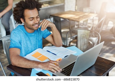 Top View Of Glad Male Student Using Laptop Computer While Writing Composition In Foreign Literature Smiling As Finding Necessary Information In Internet Sitting At Wooden Table At Cozy Cafeteria