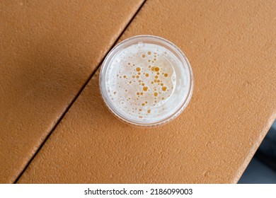 Top View Of A Glass Of Sour Flavored Beer With Its Head Covered In Froth. The Liquid Has An Orange Tint. There's A Plastic Cup Cover On The Beer On A Composite Plastic Table Top At A Microbrewery. 