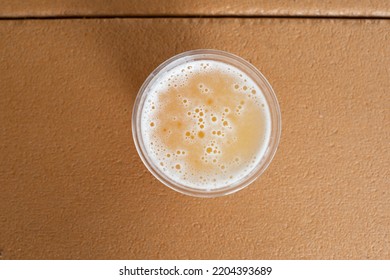 Top View Of A Glass Of Sour Flavored Beer With Its Head Covered In Froth. The Liquid Has An Orange Tint. There's A Plastic Cup Cover On The Beer On A Composite Plastic Table Top At A Microbrewery. 