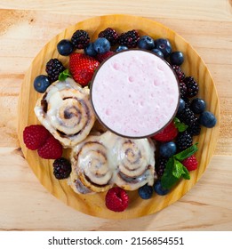 Top view of glazed cinnamon rolls served on wooden board with milk smoothie and fresh ripe berries