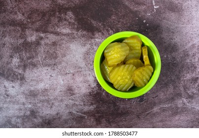 Top View Of A Green Bowl Filled With Hamburger Dill Pickle Slices Offset On A Maroon Tabletop Illuminated With Natural Lighting.