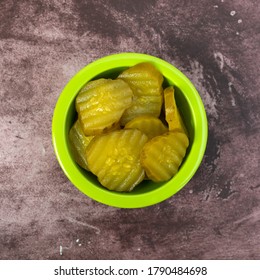 Top View Of A Green Bowl Filled With Hamburger Dill Pickle Slices On A Maroon Tabletop Illuminated With Natural Lighting.