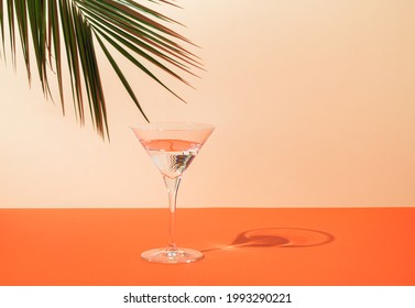 Top View Of Green Tropical Palm Leaf And Martini Glass On Orange And Sand Color Background. Summer Drink Minimal Concept. Elegant Composition.