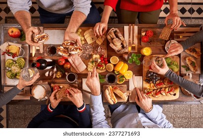 Top view group of anonymous friends eating appetizing various snacks on cutting boards while sitting at wooden table in kitchen