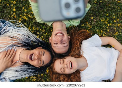 Top View Of A Group Of Cheerful Bloggers Shooting Selfie Video Reading Funny Comments On Website While Lying On A Grassy Lawn. Sunny Day And Laugh. Emotions And