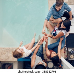 Top view of group of friends toasting at party by a swimming pool. High angle shot of young people sitting by the pool having wine. Men and women partying by the pool.