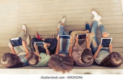 Top view of group of teenage boys and girls using tablets while sitting in row on wooden floor