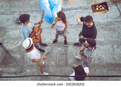 Top View Of A Group Of Young Friends Having Fun At A Rooftop Party, Playing The Guitar, Singing And Enjoying Hot Summer Days. Focus On The Couple In The Middle