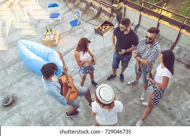 Top View Of A Group Of Young Friends Having Fun At A Rooftop Party, Playing The Guitar, Singing And Enjoying Hot Summer Days