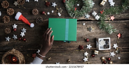 Top View Of A Hand Of Black Coloured Girl Holding A Green Holiday Gift Box On A Rustic Wooden Desk Festively Decorated With Christmas Ornaments And Decor.