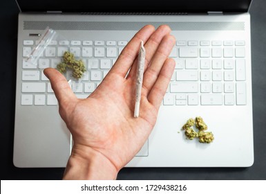 Top View Of Hand Holding A Big Marijuana Joint With Computer And Cannabis Buds On Background. Concept Of Marijuana And Technology Or Office.