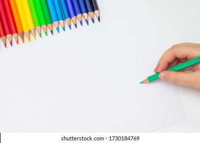 Top View Of Hand Of Kid Drawing On White Blank Of Paper With Set Of Colored Pencils. Copy Space For Text. Mockup.