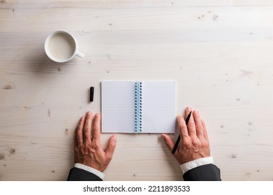 Top View Of Hands Of A Businessman Holding Black Marker By An Open Spiral Notebook On Wooden Desk With A Cup Of Coffee, 