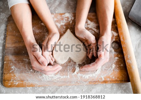 Top view, hands close-up. Mom and daughter make cookies in the form of Wednesday, together roll out the heart with a rolling pin on a wooden board in flour