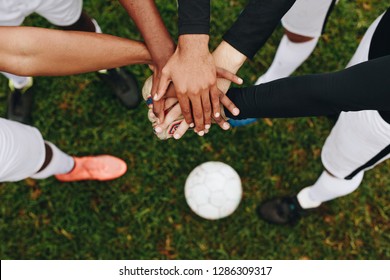 Top view of hands of players placed one over the other standing in a huddle. Players standing in a huddle joining their hands together in the centre with a soccer ball on the ground. - Powered by Shutterstock