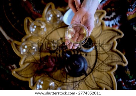 Top view of hands presenting a clear teacup over a floral tea tray with cups and teapot during a quiet indoor ritual. Tea meditation and slow routines help reduce stress and restore emotional balance.