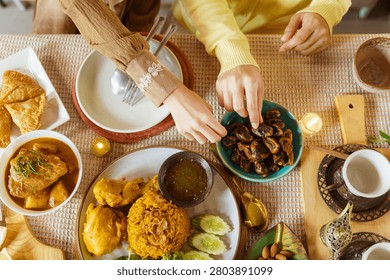 Top view of hands reaching for dates on a festive iftar table spread with spiced rice, curry chicken, samosa, dipping sauce, almonds and candles for Ramadan celebration.