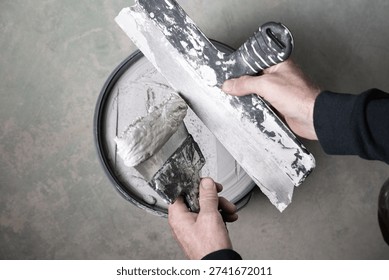 Top view of hands transferring wall filler from a putty knife onto a wide taping knife above a bucket. Concept of drywall finishing, wall repair and DIY home renovation work.